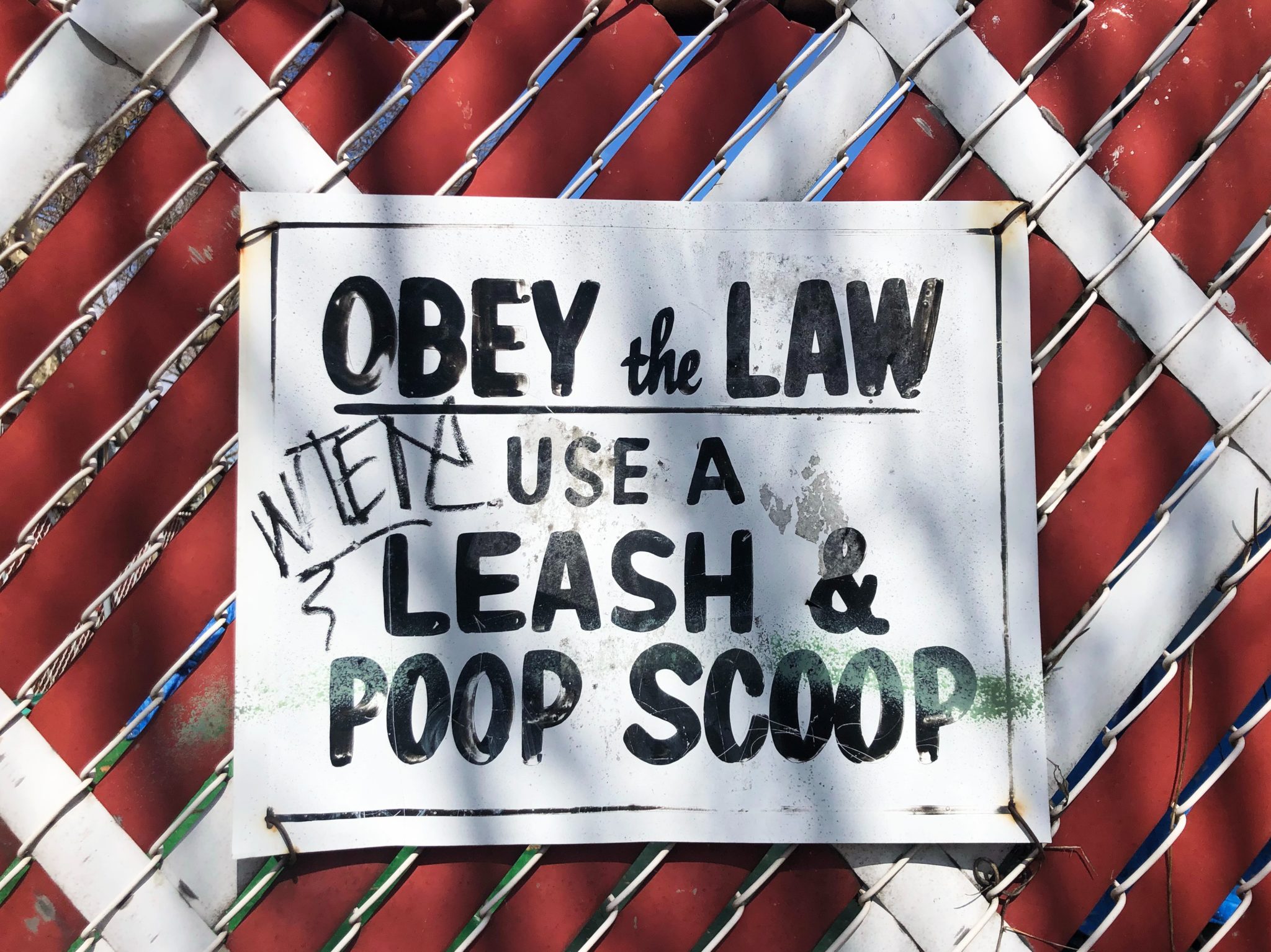 Photo of white metal sign with black text that says 'Obey the Law - Use a Leash and Poop Scoop' attached to a chainlink fence in front of a red wall